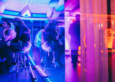 People sitting behind a table, bathed in sharp blue light, and other people reflected in a surface showing an orange light.