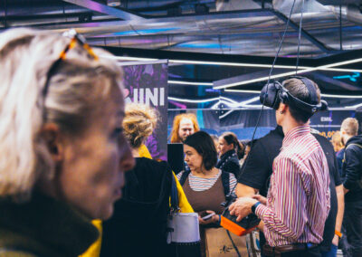 A blurry person in the foreground, with another person using a VR headset in the background, at an event.