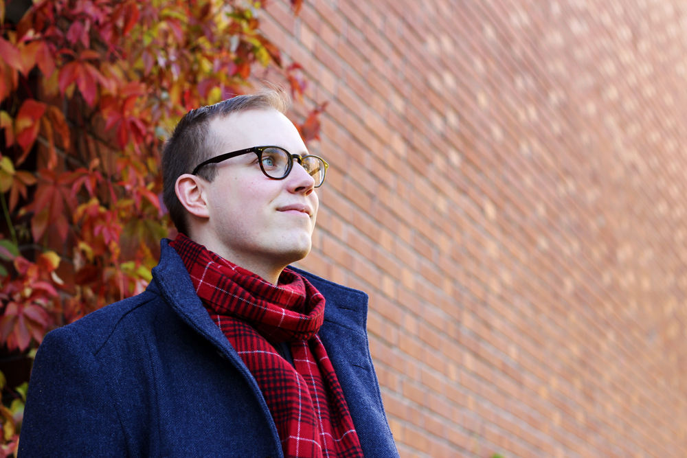 Man in a blue coat and red scarf standing in front of a brick wall with red ivy.